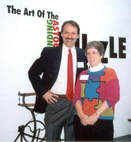 Image of Will Shortz and Anne Williams standing in front of museum exhibit of jigsaw and mechanical puzzles